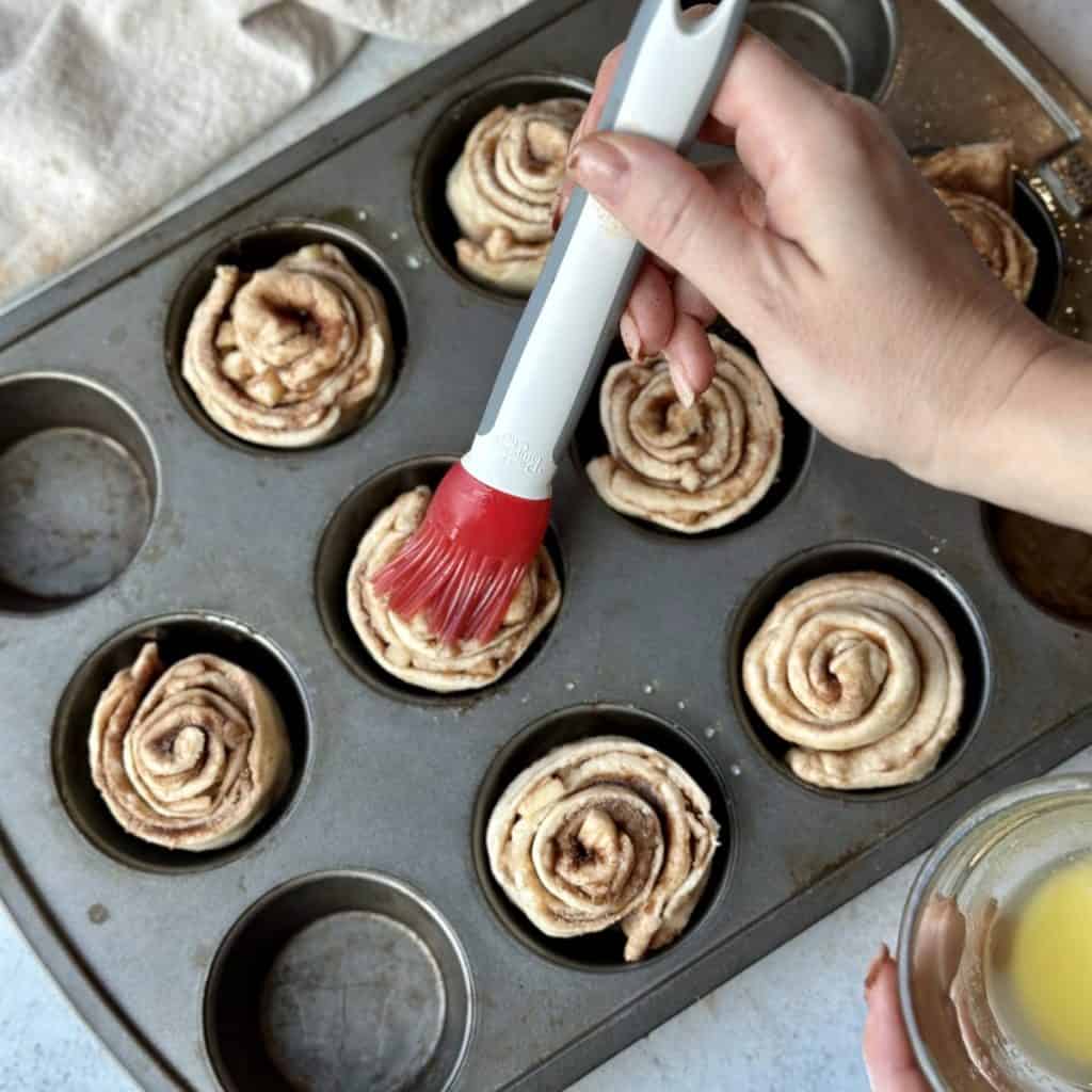 Brushing butter on butter on cinnamon rolls in a muffin pan.