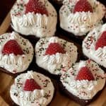 This is a picture of brownies with a cream cheese frosting and Santa hat on top made with a halved strawberry and whipped cream.