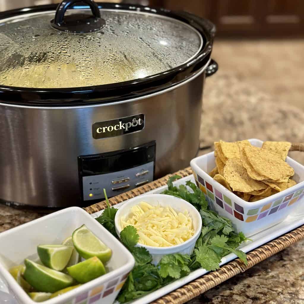 a crockpot that has green chicken enchilada soup inside with the toppings, lime wedges, shredded cheese, chips and cilantro on a serving tray