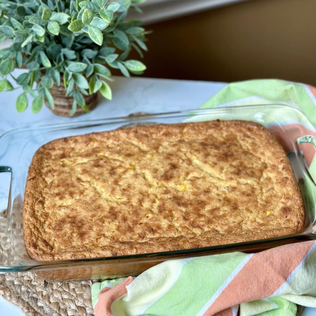 Country scalloped corn in a baking dish with a green and orange dish towel around it.