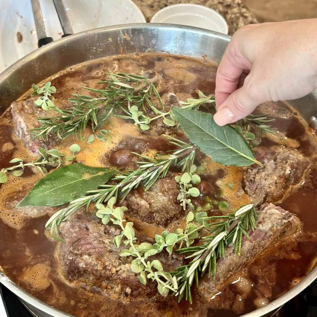 Adding fresh herbs to the top of beef short ribs and broth in a pan.