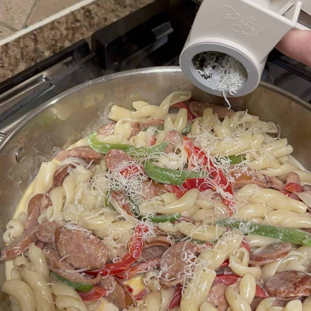 Adding parmesan to a skillet of noodles, vegetables and sausage.