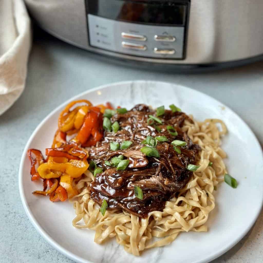 A plate of slow cooker Korean beef, noodles and vegetables next to a crockpot.