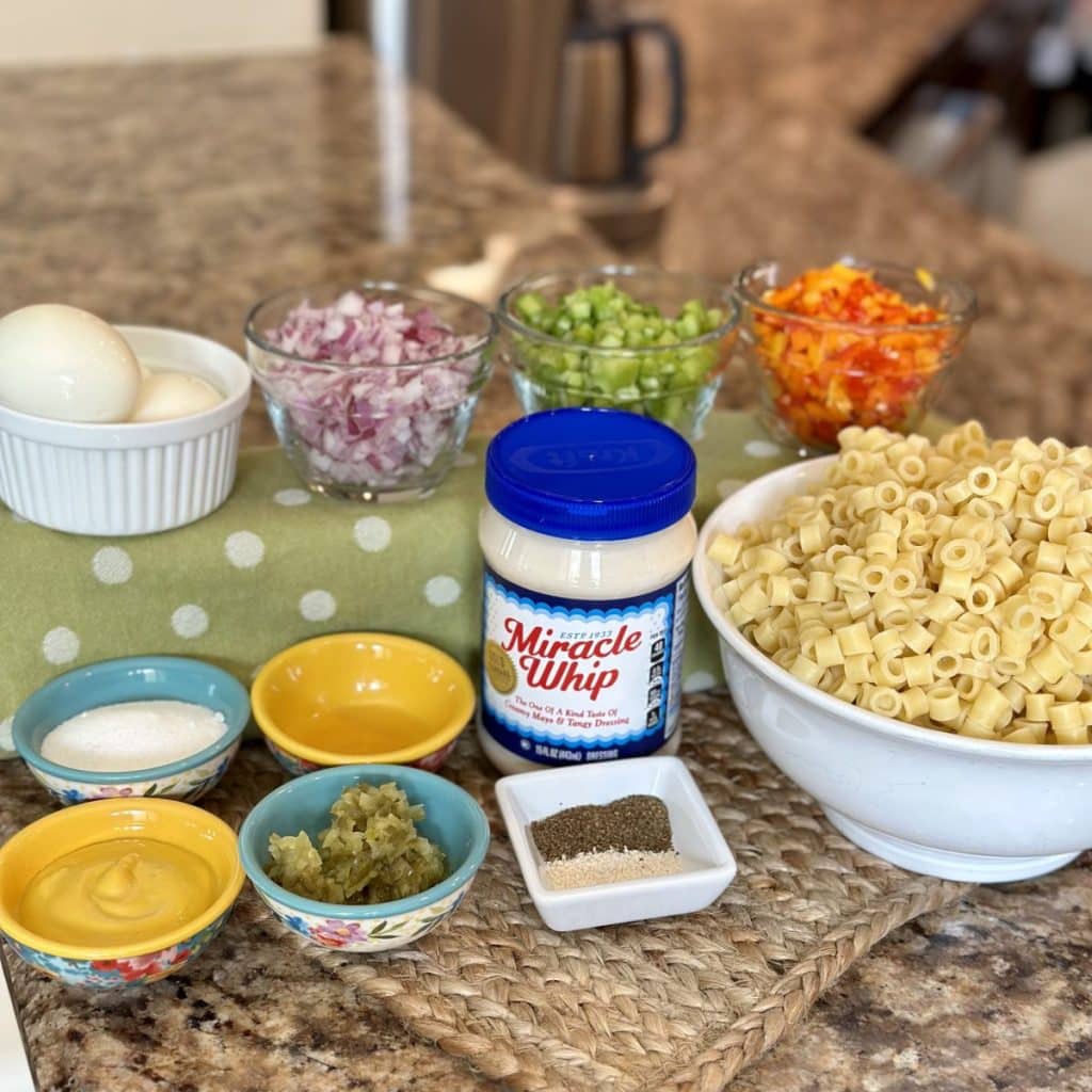 Ingredients to make macaroni salad: miracle whip, sugar, celery seed, garlic salt, apple cider vinegar, dill pickle relish, yellow mustard, pasta, eggs, red onion, celery and bell peppers.