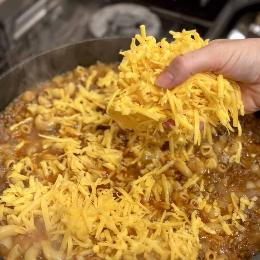 Adding cheddar cheese to a skillet of beef and broth with noodles.