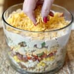 Adding shredded cheese to the top of a cornbread chicken salad in a trifle bowl.