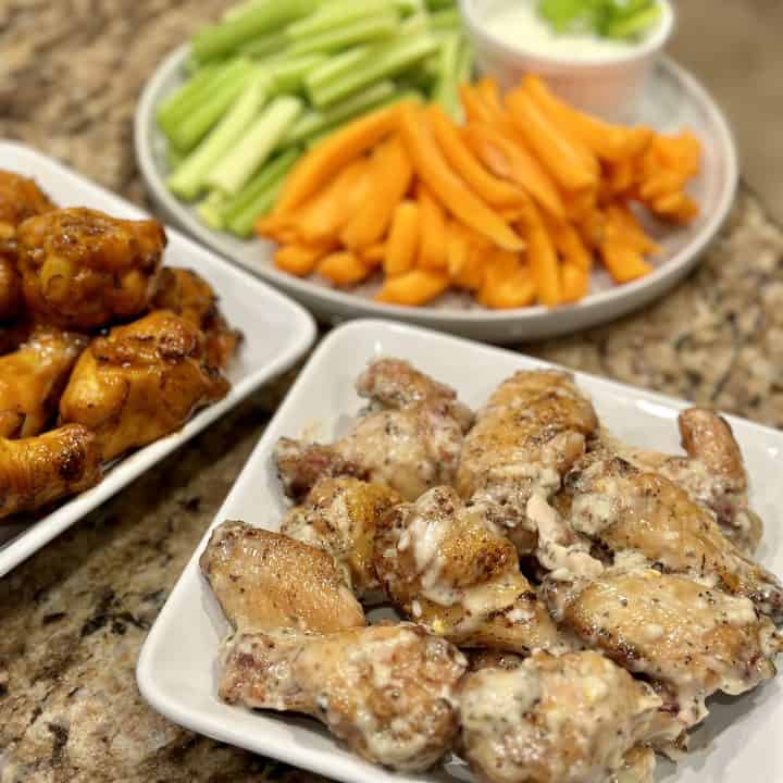 This is a picture of chicken wings that have been tossed in a light brown garlic sauce. There is plate of cut up carrots and celery with dip.
