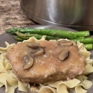 A plate topped with noodles. On top of the noodles is a pork chop with brown gravy. Next to the pork chop is green asparagus.