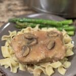 This a picture of a pork chop on top of yellow noodles with brown gravy with mushrooms poured on top. Green asparagus is laying next to it.