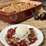This is a picture of a saucer with a cherry dessert on top. The cherry is on the bottom in a glaze filling and there is an oatmeal brown sugar crumble on top. The dessert is topped with a scoop of vanilla ice cream.