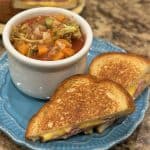 This is a picture of a bowl of garden vegetable soup and a sandwich cut in 2 pieces. The bowl of soup is filled with red tomato juice, orange carrots, green broccoli and celery and white cauliflower. The sandwich has yellow cheese and ham between the slices of bread.