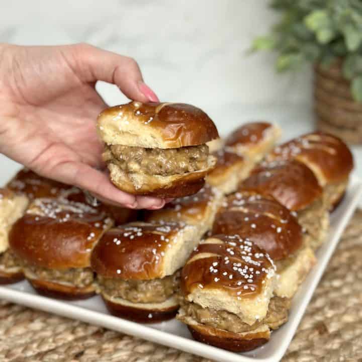 This is a picture of a miniature sandwich. Between the pretzel bun is a filling of yellow cheese and hamburger and sausage meat. There’s also a tray of these sandwiches in the background.