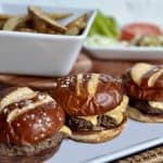 This is a picture of cheeseburgers. They’re on a brown pretzel bun on a white platter. Behind the cheeseburger is a bowl of potato wedges and toppings for the burgers that includes tomatoes, green leaf lettuce, chopped onions and pickles.