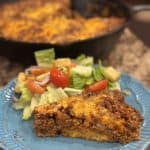 This is a picture of a slice of meat pie. The bottom level is cornbread and top is a mixture of beef, onion and manwhich tomato sauce. It’s topped with cheese. Next to is is a tossed green garden salad with tomatoes.