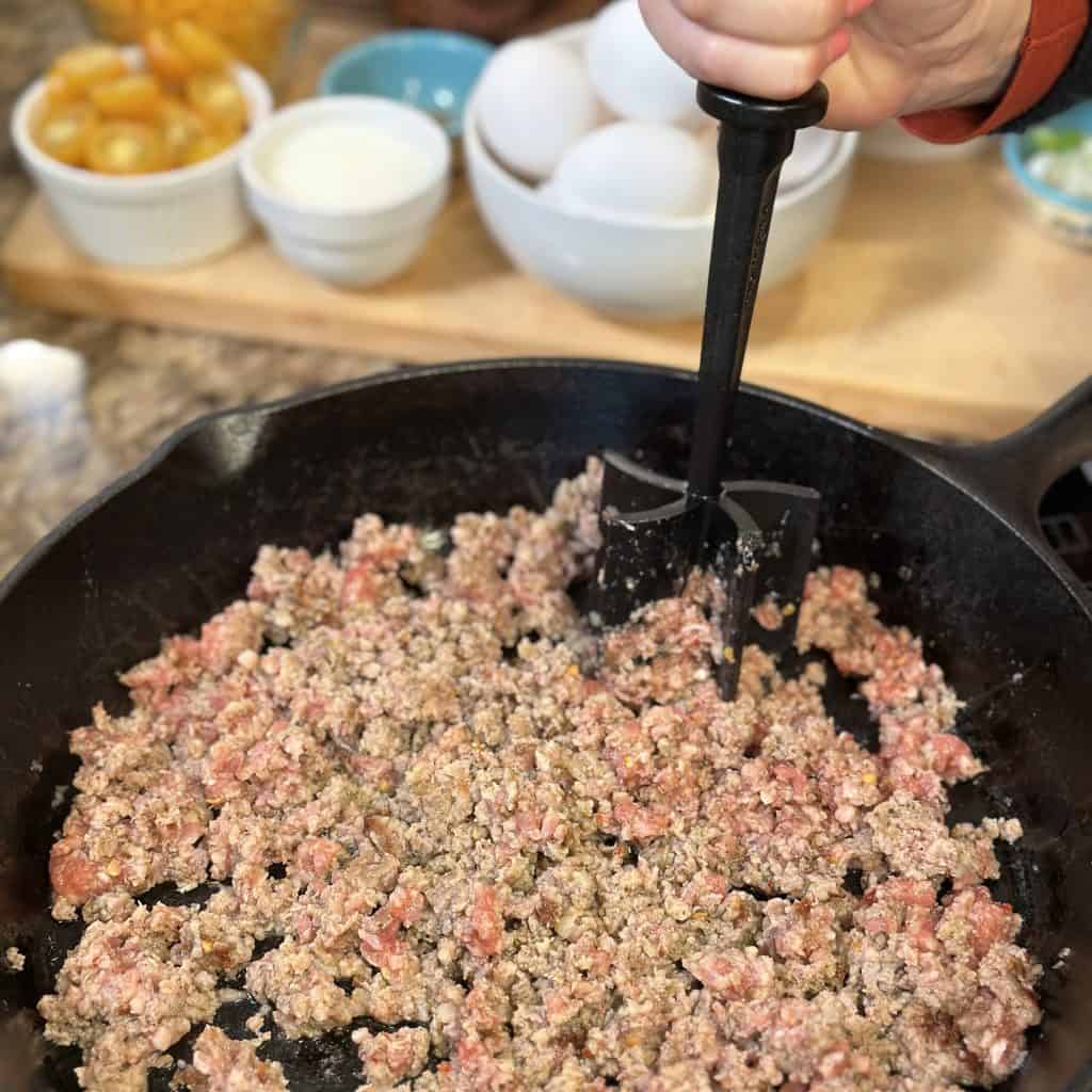 breaking up cooking sausage in a cast iron skillet with a meat chopper