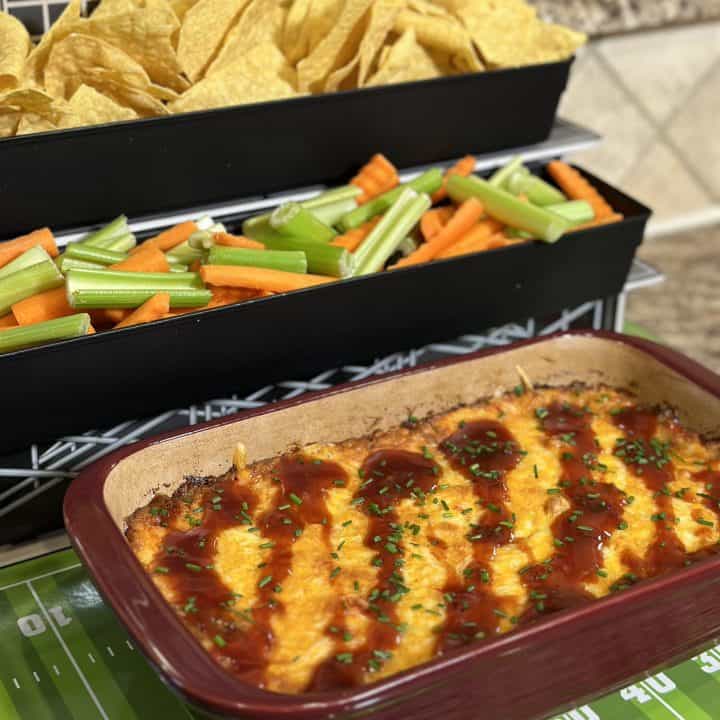 This is a picture os a maroon baking dish filled with a yellow cheese dip with barbecue sauce on top. There is also orange carrots and green celery next to it as well as chips.