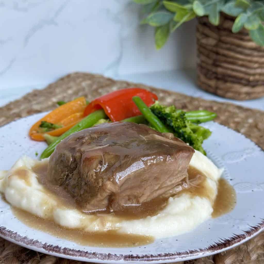 This is a picture of a plate topped with mashed potatoes and a slice of tender pork. There is brown gravy on top of the pork. Next to the potatoes and pork are some steamed green beans and sweet peppers.
