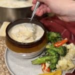 This is a picture of a brown bowl full of a cream based chicken noodle soup. Next to the bowl on a plate is a cooked veggie blend of cauliflower, peppers and broccoli.