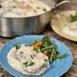This is a picture a blue plate with mashed potatoes on top. On the mashed potatoes rests a piece of chicken with a white garlic parmesan sauce and sprinkled with parsley. Next to the chicken is cooked green beans and broccoli.