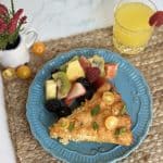 This is a picture of a slice of breakfast casserole on a blue plate with a side of a fruit salad with kiwi, pineapple, raspberries and orange slices. The casserole has tater tots and cheese on top and is garnished with tomatoes. There is also a glass of orange juice in the picture and a purple flower.