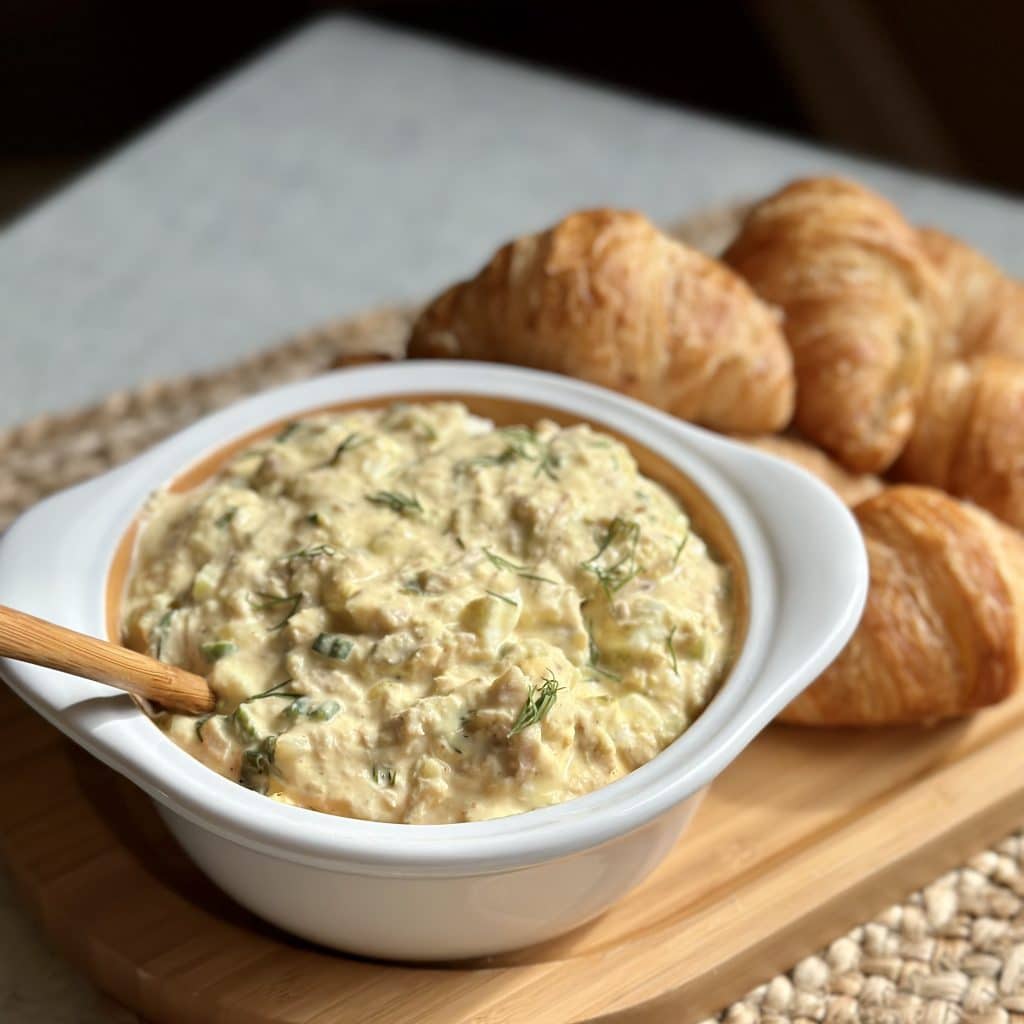 A bowl of yellow egg salad with green dill on top. Croissants are beside the bowl
