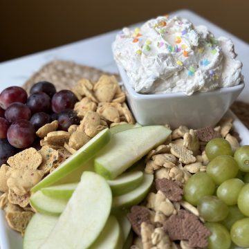 This is a picture os a bowl of white fruit and snack dip. Next to the dip is red and green grapes, rabbit shaped cookies and a sliced green apple. There are sprinkles on top of the dip.