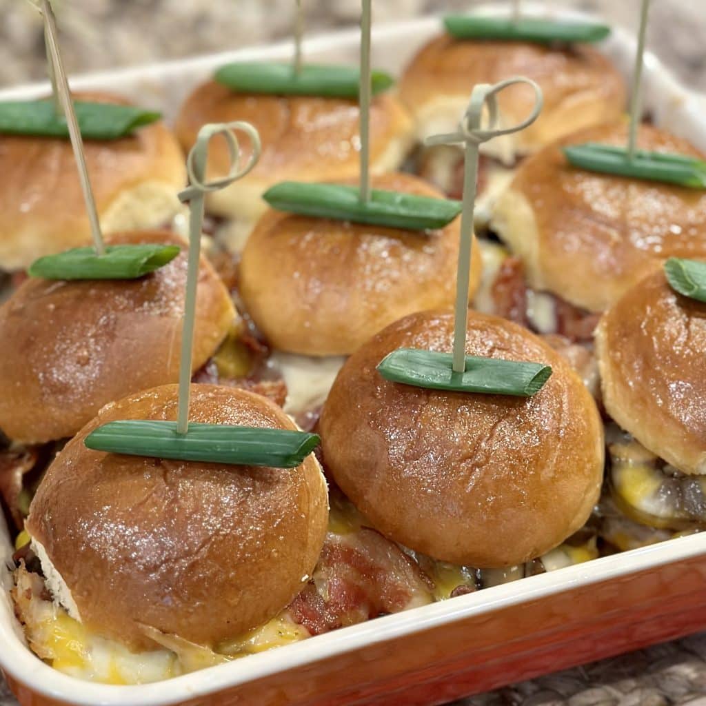This is a picture of a pan of mini sandwiches. The filling is chicken, bacon and melted cheese. The sandwich has a skewer holding it together an it’s garnished with a green onion.