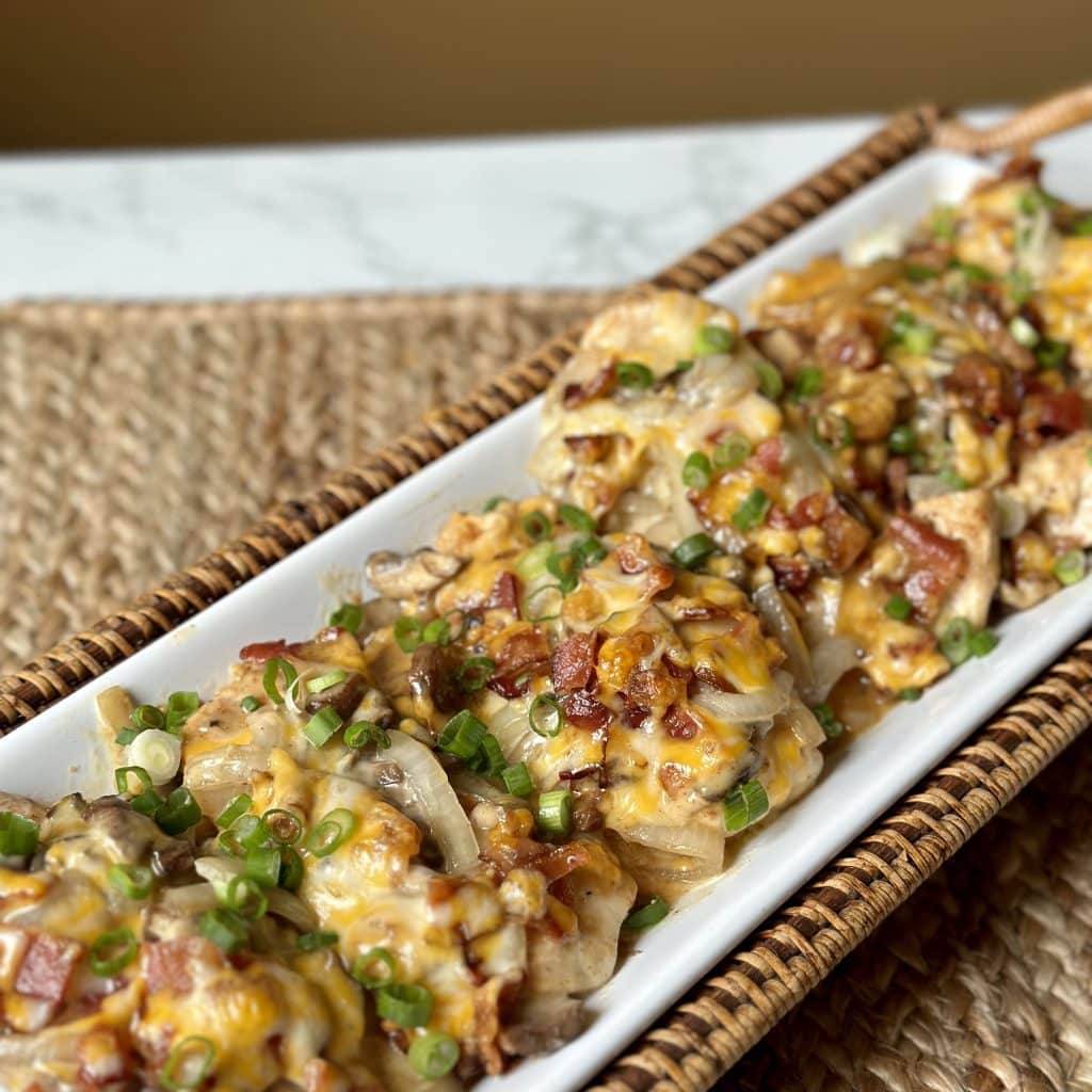 This is a picture of a plate of chicken. On top of the cooked chicken is crumbled bacon, green onions, melted yellow cheese and cooked mushrooms and onions