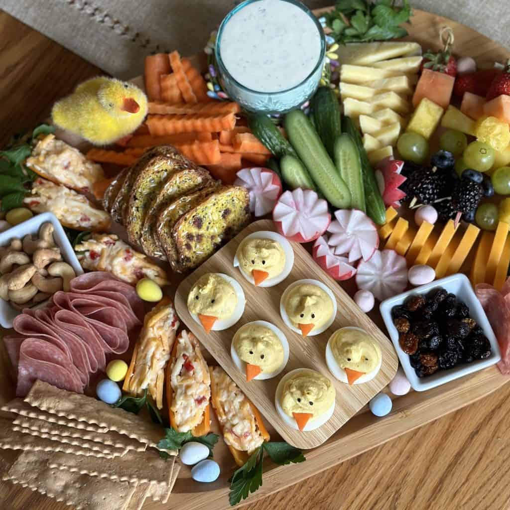 A charcuterie board with crackers, cheddar cheeses, deviled eggs, a rainbow colored fruit assortment, raw veggies, a ranch dip, mixed nuts, dried fruit and pimento cheese stuffed carrots