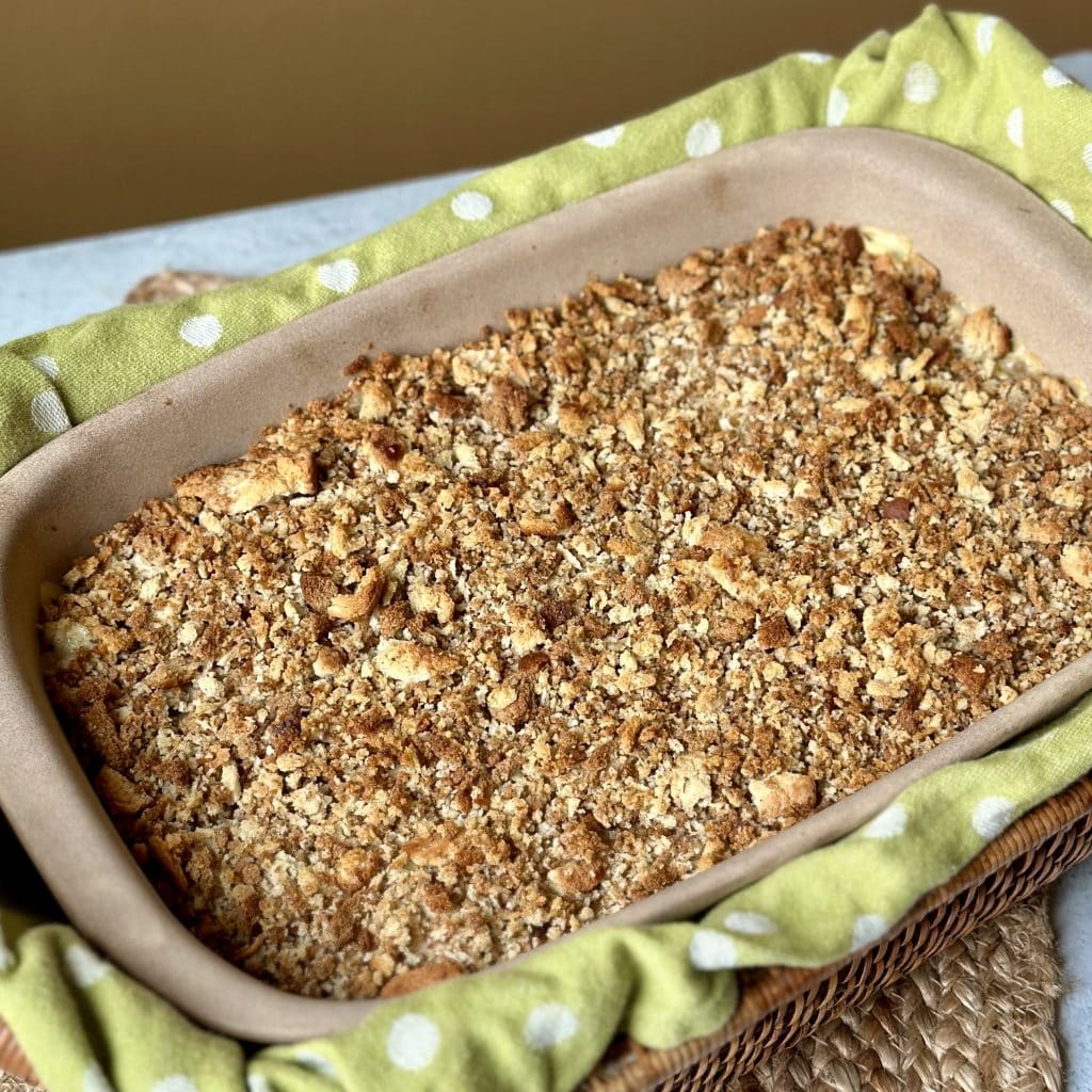 This is a picture of a chicken casserole dish. The top is golden brown stuffing and below the stuffing are layers of chicken, cheese and a chicken gravy. The dish is laying in a basket with a green towel.