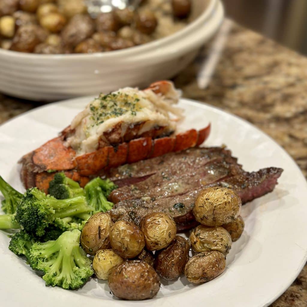 This is a picture of a plate of food. On the plate is green broccoli, a garlic butter seasoned lobster tail, sliced steak and roasted baby potatoes.
