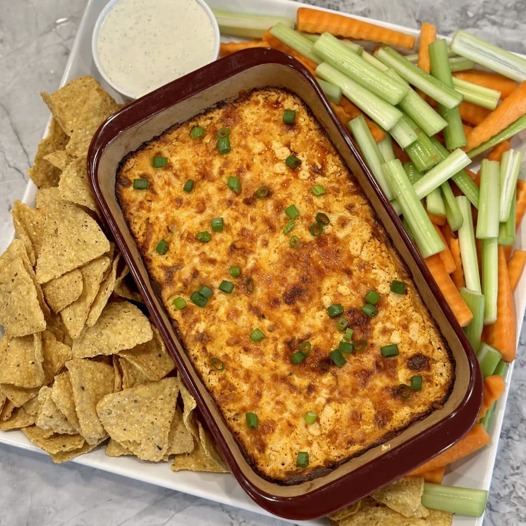 Baked cheesy chicken dip in a glass pan surrounded by tortilla chips and celery sticks