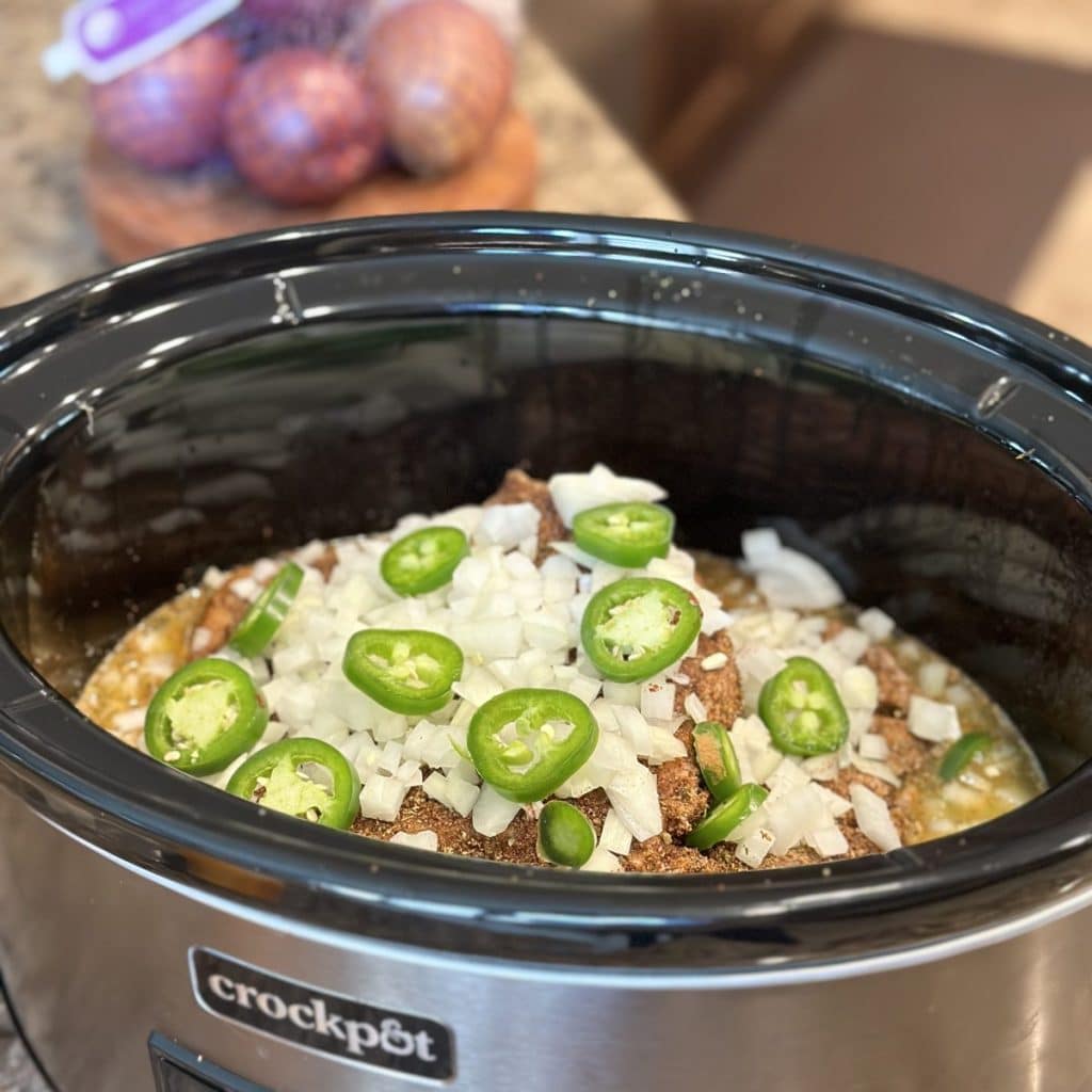 A pork shoulder in a crockpot with seasonings, onion and jalapeño chopped on top.