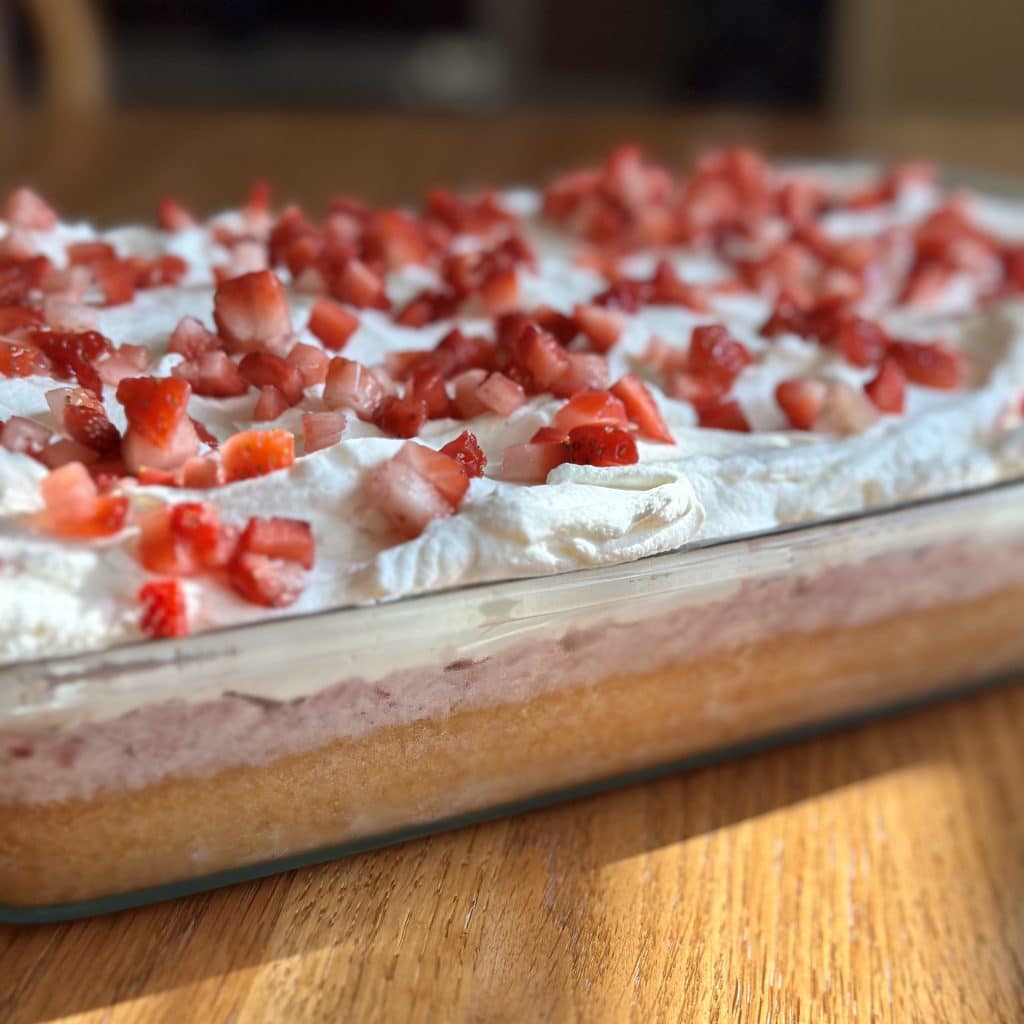 A close up shot of a cake showing 3 layers: the cake, the milk topping and the whipped topping with chopped strawberries on top