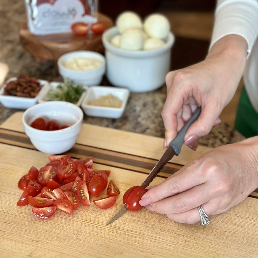 Grape tomatoes being quartered on a cutting board, with deluxe BLT deviled egg ingredients displayed in background