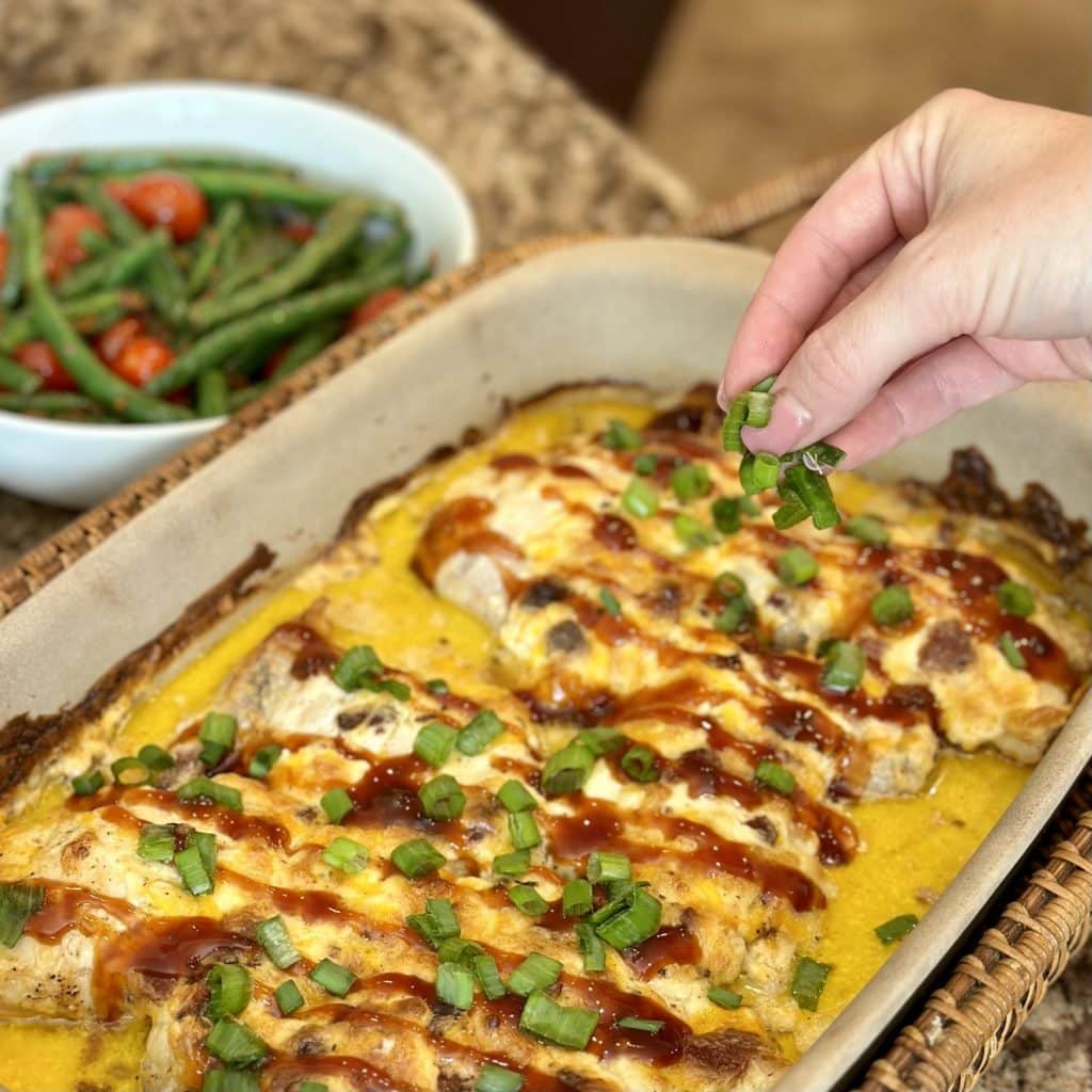 A pan of cooked chicken with barbecue sauce on top and being sprinkled with green onion slices