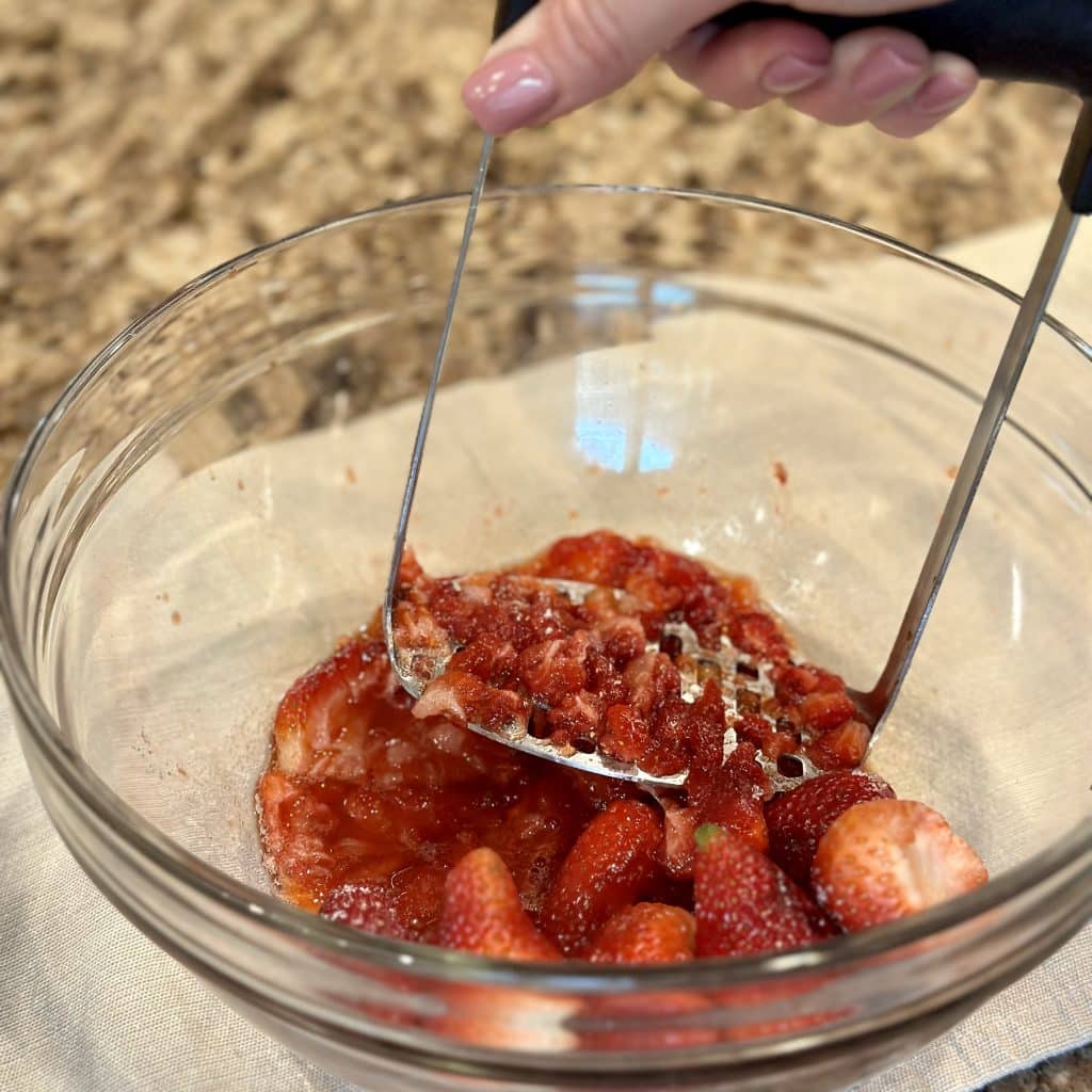 A potato masher mashing together strawberries and sugar in a glass bowl