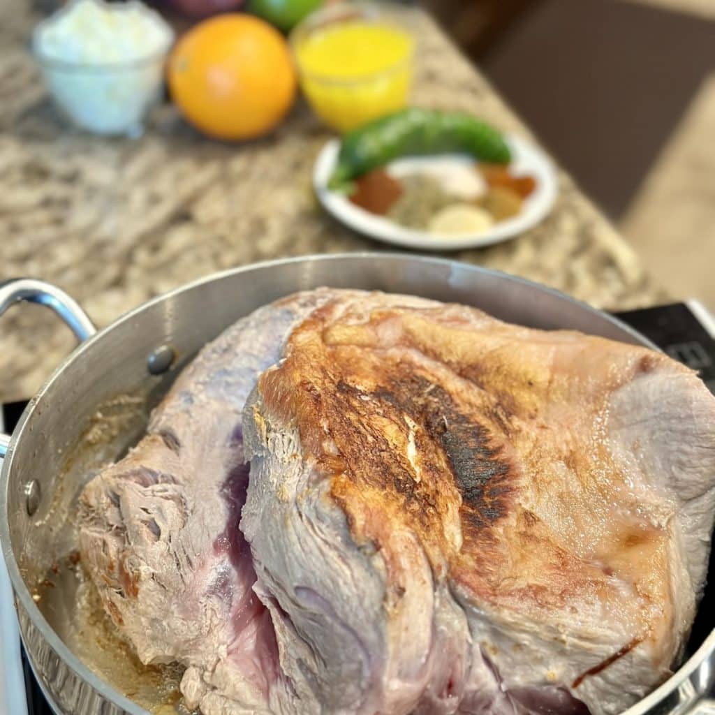 A pork shoulder searing in oil in a skillet.