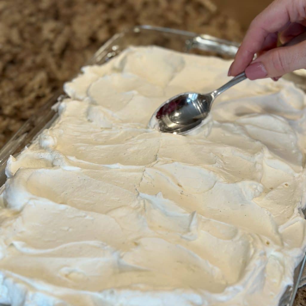 A spoon spreading whipped topping on the top of a vanilla cake