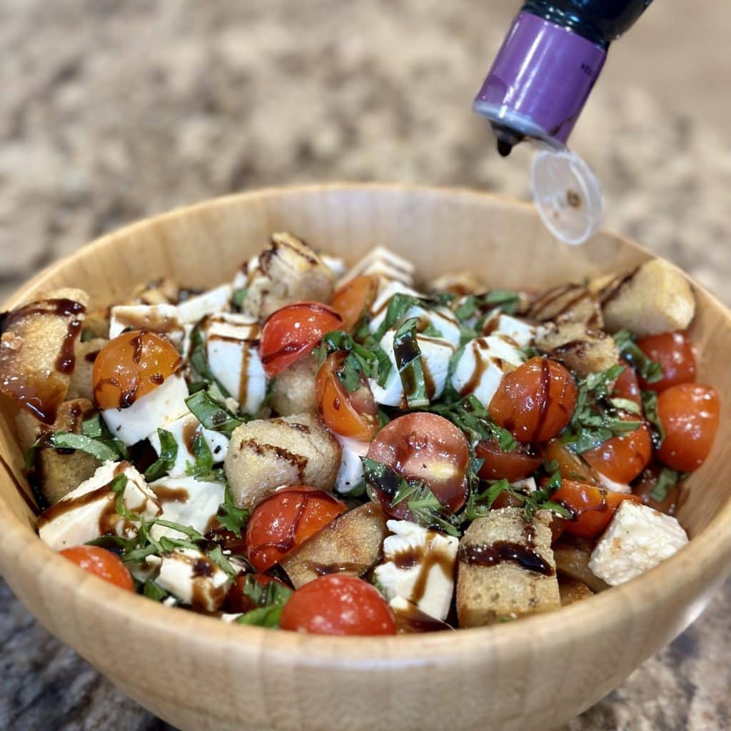 A bowl filled with a mixture of tomatoes, mozzarella, basil, and bread cubes and being drizzled with balsamic glaze.