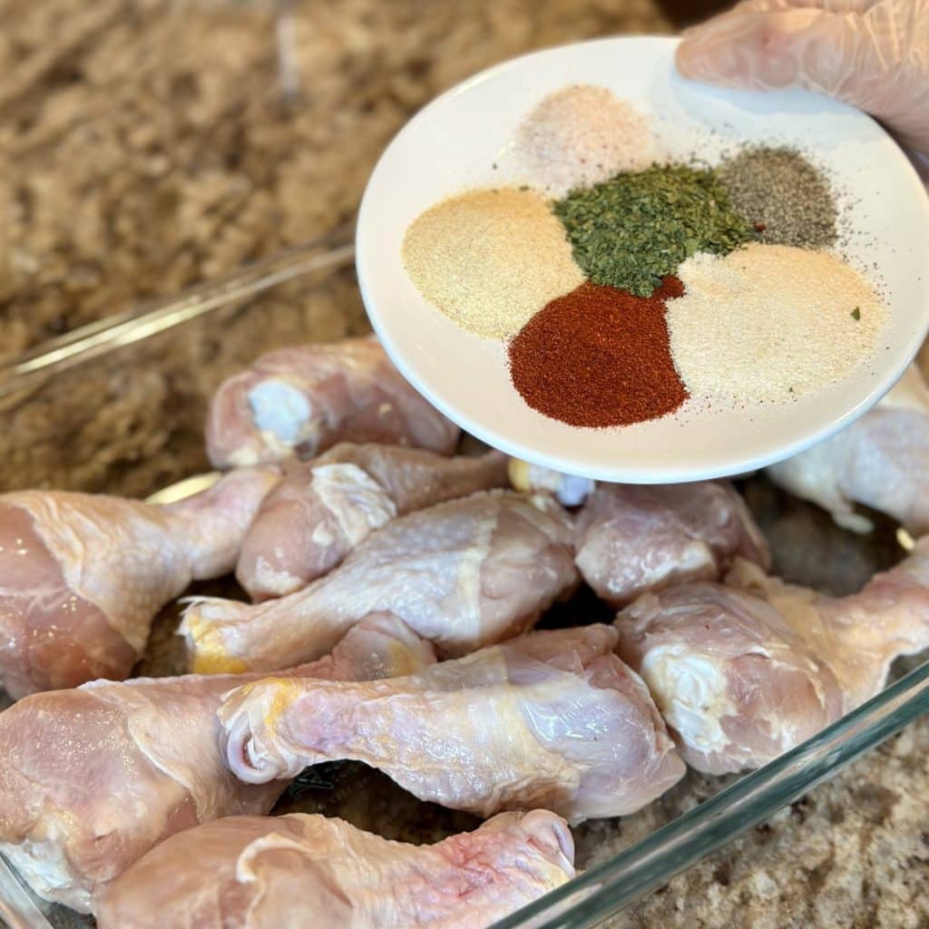 Seasonings being added to drumsticks in a baking dish.