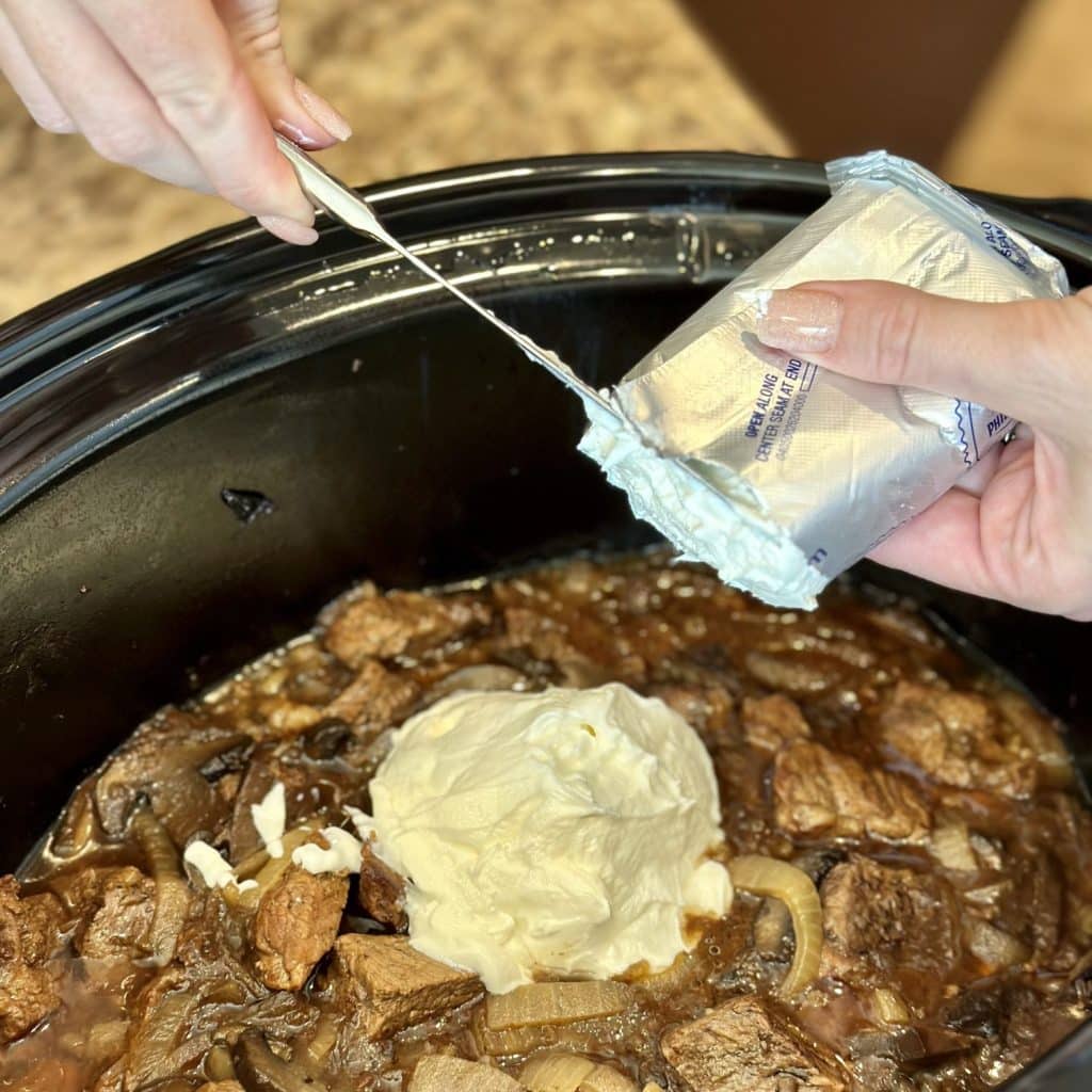 Cream cheese and sour cream being added to a crockpot of beef stroganoff.