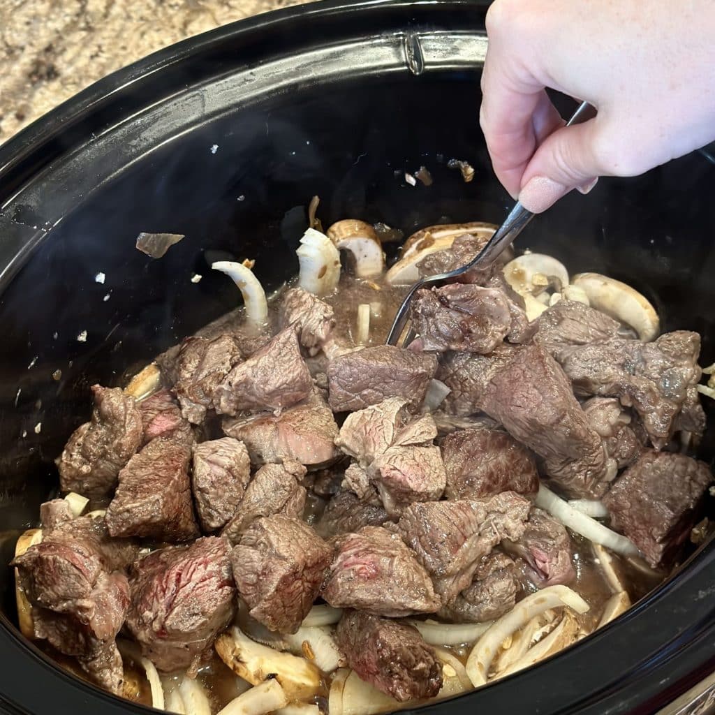 Mixing seared cubed beef in a crockpot with veggies and French onion soup.