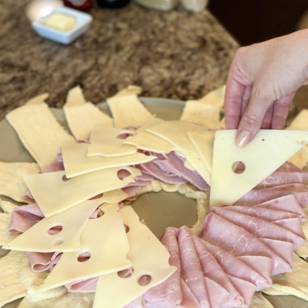 One hand placing swiss cheese triangles on top of a crescent roll ring layered with ham.