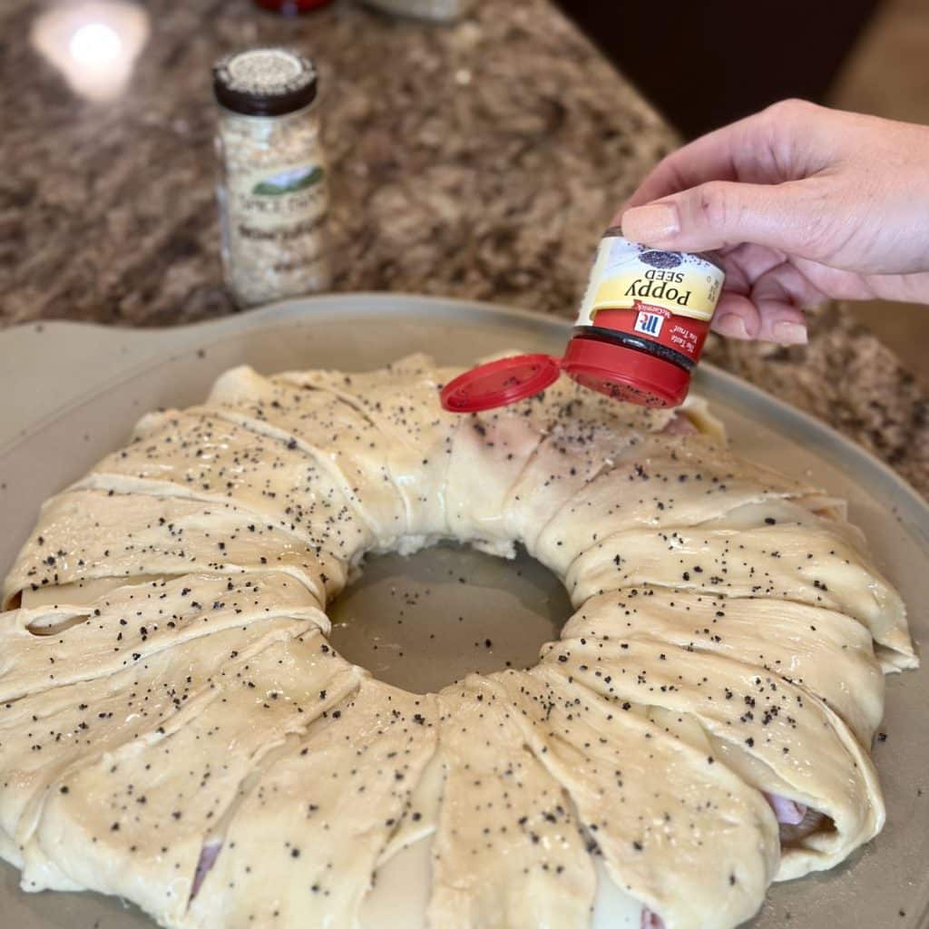 One hand sprinkling poppy seeds on top of crescent ring with spices in the background.