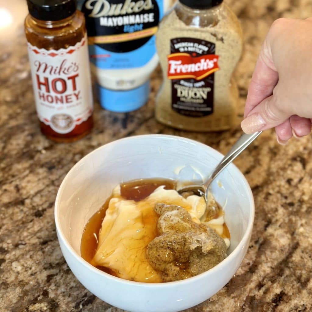 One hand using a spoon to stir together in a small bowl mayonnaise, honey and dijon mustard. The ingredient bottles are shown in the background. 