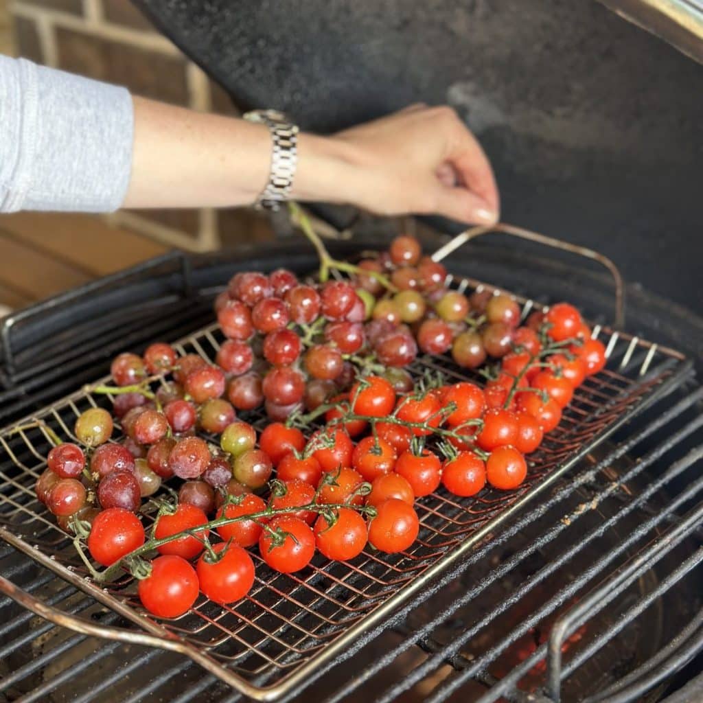 Placing a grill basket of tomatoes and grapes on a grill.