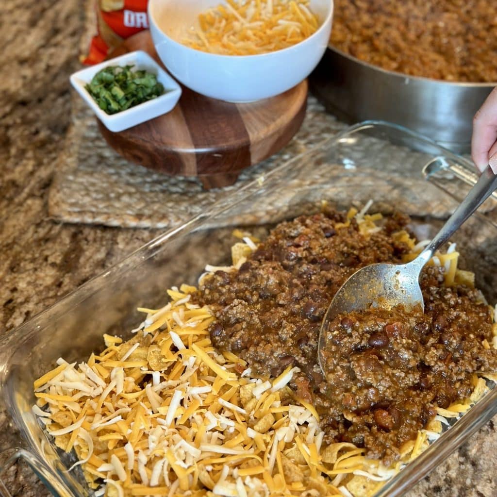Spreading cooked ground beef on Fritos and cheese in a baking dish.