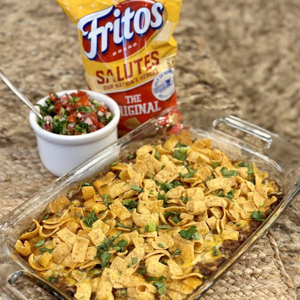 A baking dish full of Frito taco pie with a bag of Fritos and pico de gallo on the side.