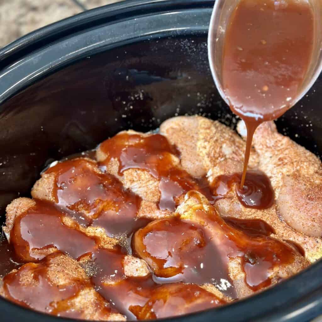 Adding barbecue sauce to the top of chicken legs in a crockpot.